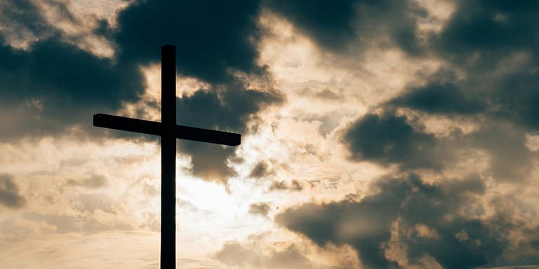 Image of sun breaking through dark clouds with a cross silhouetted against the sky